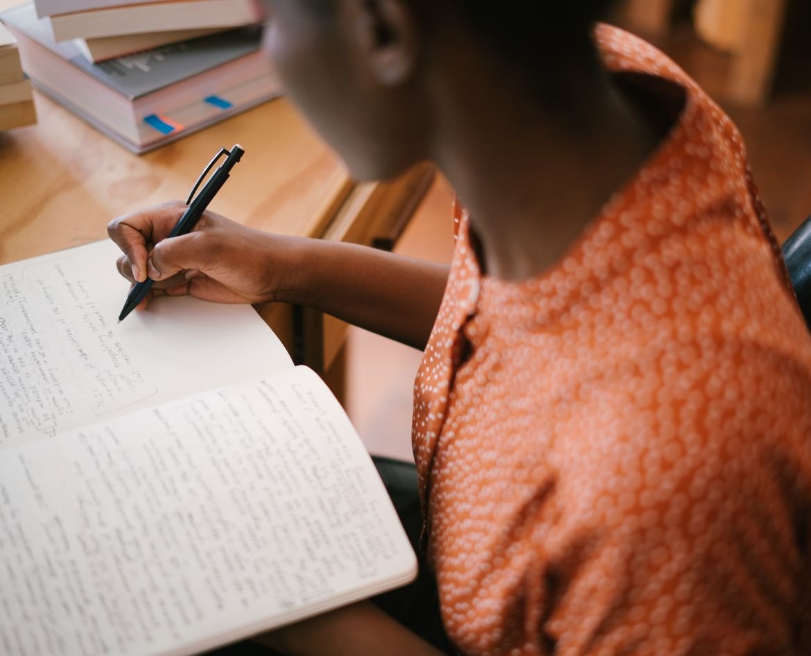 Student writing study notes in a notebook beside a stack of books